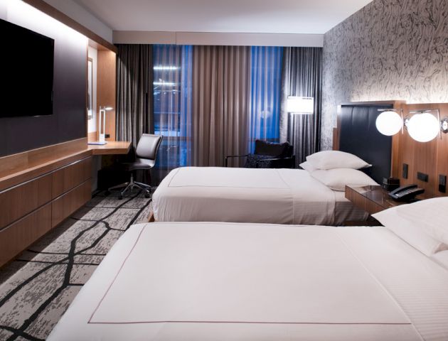 A modern hotel room with two beds, wall-mounted TV, desk, chair, and large windows with curtains, featuring a stylish design and ample lighting.