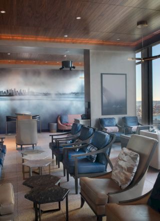 A modern lounge room with comfortable seating, large windows offering a waterfront view, and a wall mural depicting a city skyline.