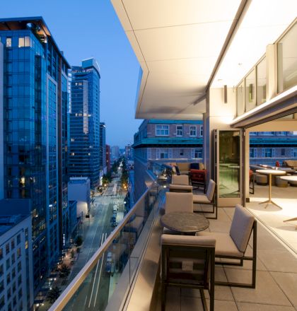 A modern cityscape at dusk from a rooftop terrace with seating, tables, and large windows, overlooking high-rise buildings and street view.