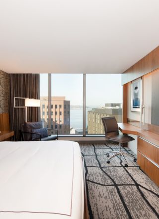 A modern hotel room with a large bed, desk, chair, flat-screen TV, and a window offering a city view.