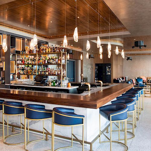 A modern bar with a wooden counter, blue stools, and hanging lights. Numerous bottles line the shelves, and there's seating in the background.