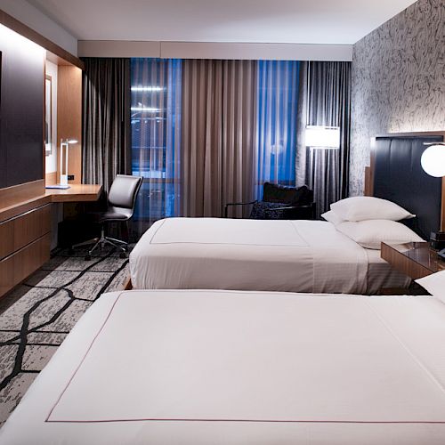 A modern hotel room with two beds, a wall-mounted TV, a desk with a chair, bedside tables with lamps, and patterned wallpaper.