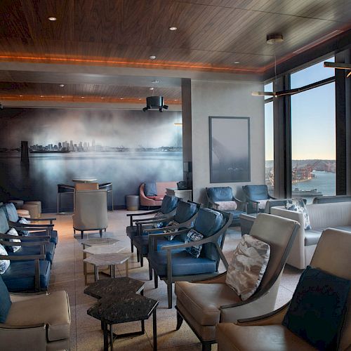 A modern lounge area with multiple chairs and tables, large windows with a waterfront view, and a mural on the back wall depicting a cityscape.