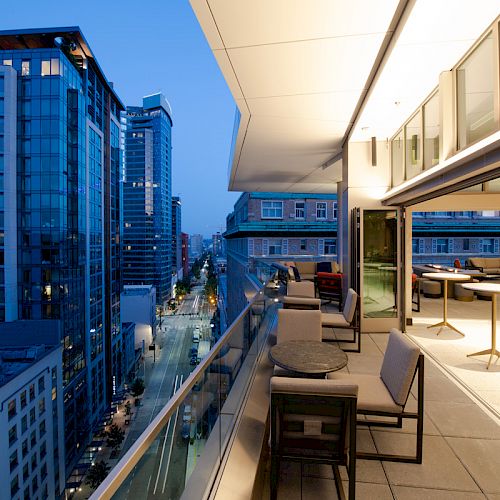 A modern cityscape view from a high-rise balcony in the evening, featuring outdoor seating and tall buildings lining a street.