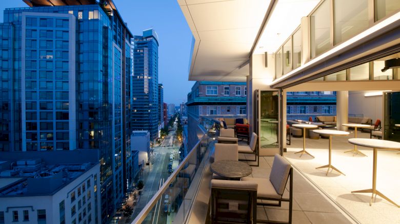 Cityscape view from a modern balcony at dusk, featuring tall buildings, outdoor seating, and a well-lit indoor area.