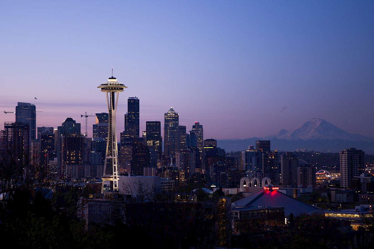 The image features the Seattle skyline at dusk, including the Space Needle and Mount Rainier in the distance, against a clear evening sky.