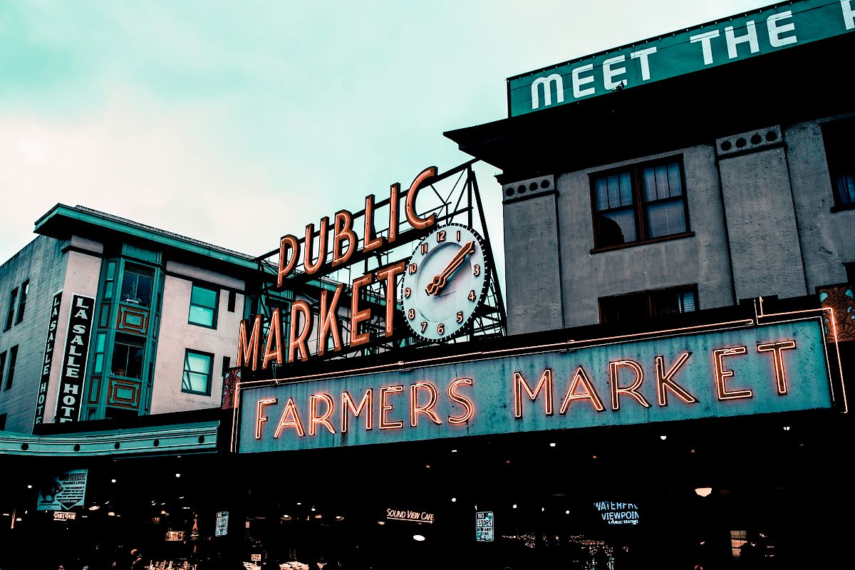 The image shows the iconic neon sign of a public market with a clock, alongside "Farmers Market" signage, situated between buildings.