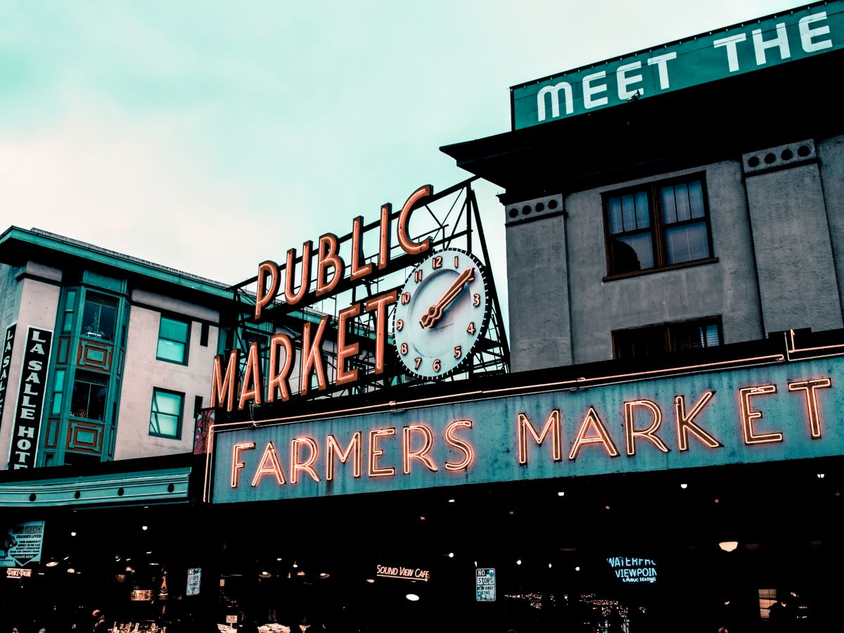The image shows the iconic neon sign of a public market with a clock, alongside "Farmers Market" signage, situated between buildings.