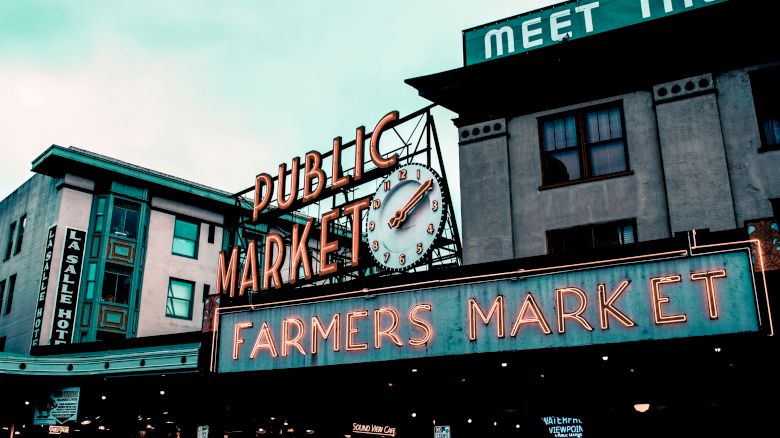 The image shows the iconic neon sign of a public market with a clock, alongside "Farmers Market" signage, situated between buildings.