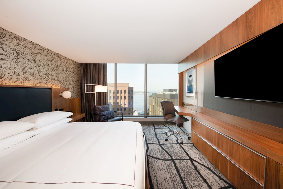 A modern hotel room with a large bed, wall-mounted TV, desk, chair, and floor-to-ceiling windows offering a city and waterfront view.