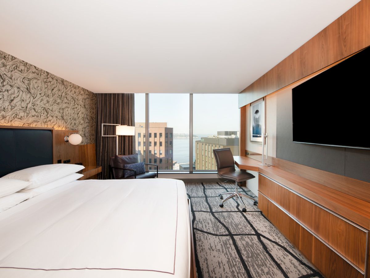 A modern hotel room with a large bed, wall-mounted TV, desk, chair, and floor-to-ceiling windows offering a city and waterfront view.