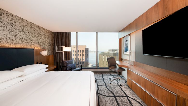 A modern hotel room with a large bed, wall-mounted TV, desk, chair, and floor-to-ceiling windows offering a city and waterfront view.