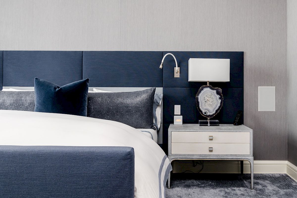 The image shows a modern bedroom with a blue and white bed, a nightstand with a lamp, and a wall-mounted reading light.