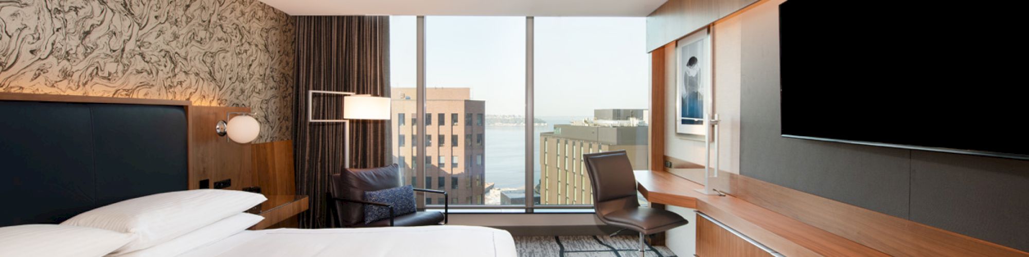A modern hotel room with a large bed, desk, chair, TV, and floor-to-ceiling windows offering a city and water view.