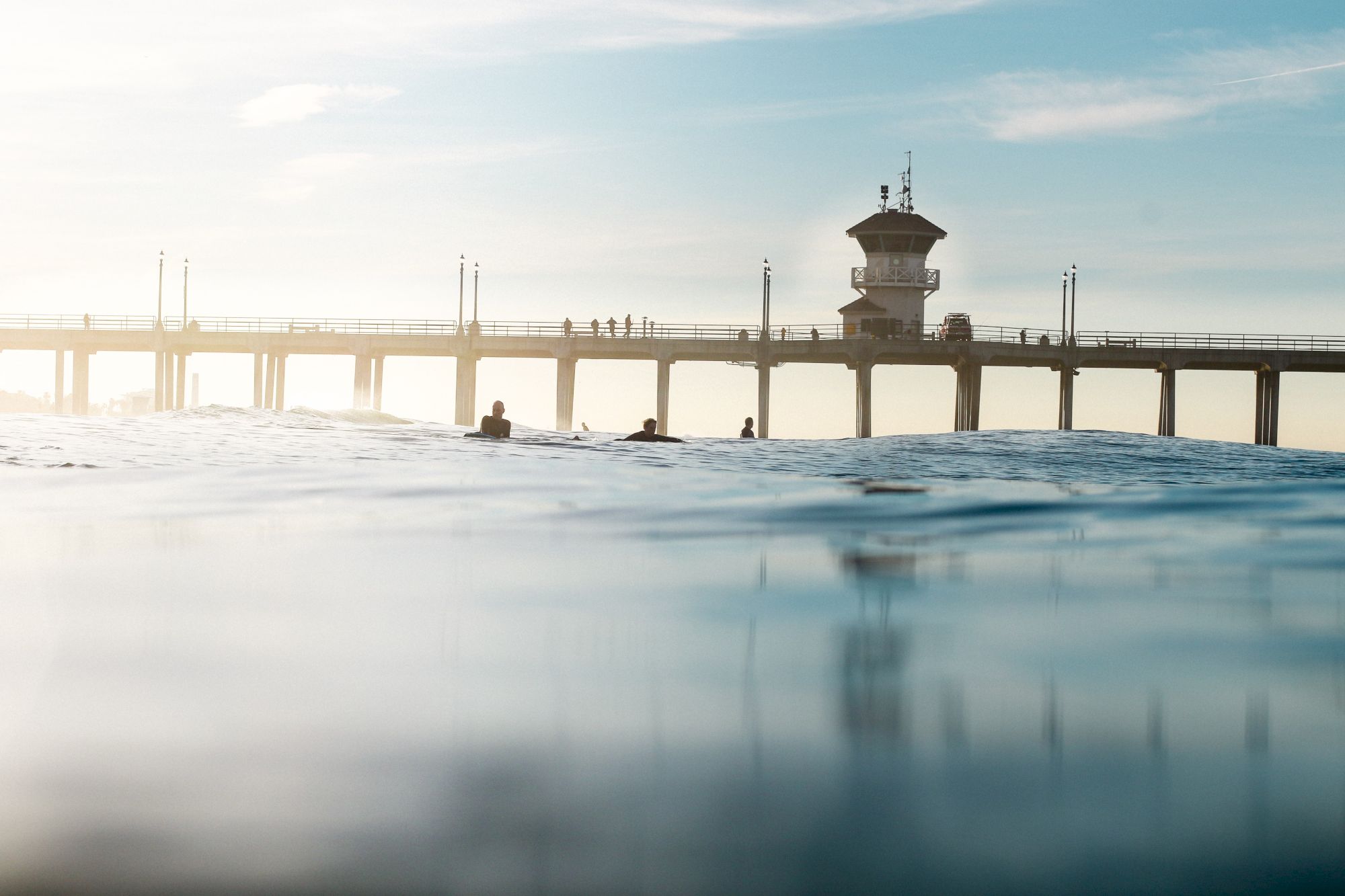 The image shows a seaside pier stretching over the water, with people on the pier and some surfers in the ocean below, under a blue sky.